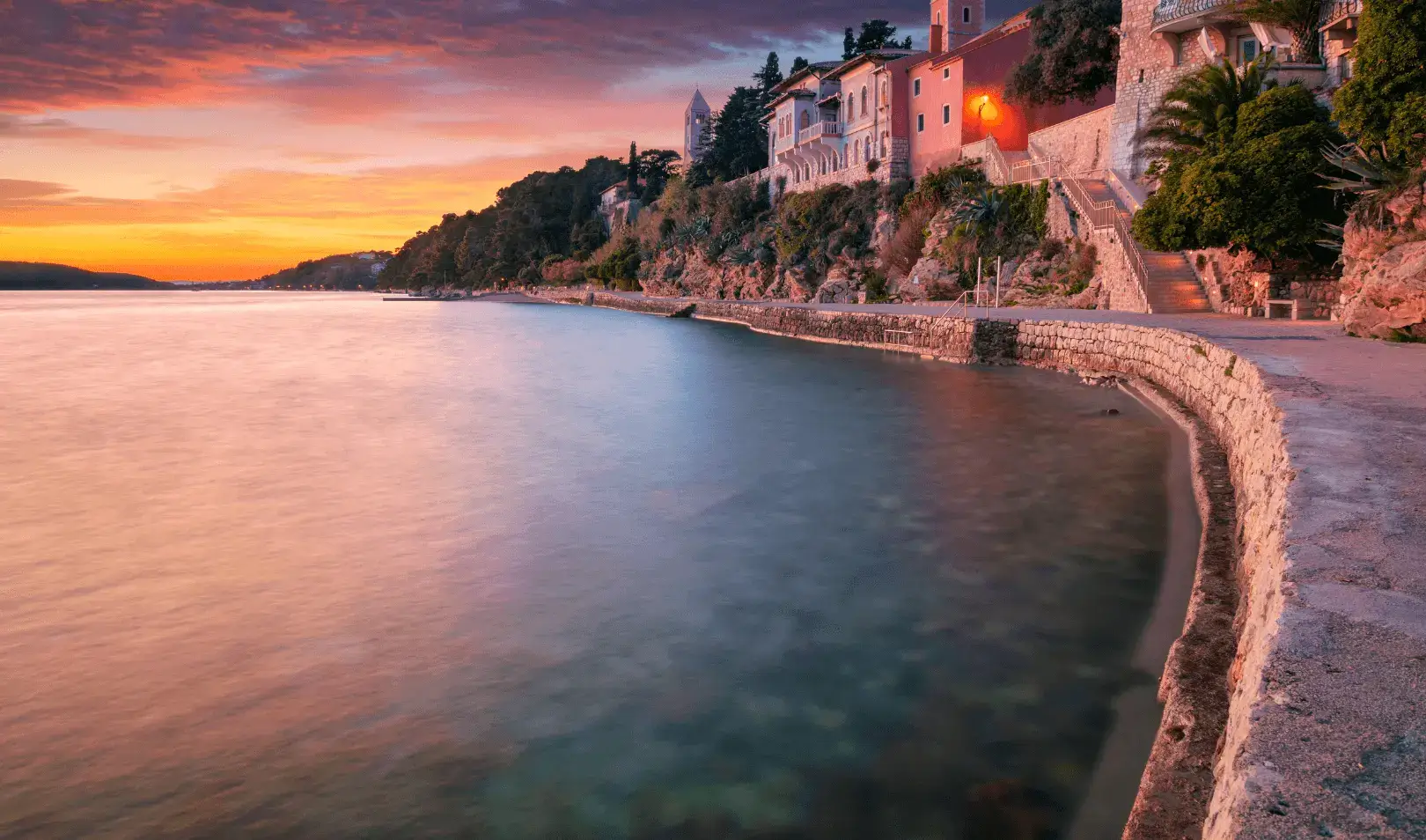 Sunset over a coastal promenade with stone houses on a hillside